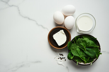Obraz na płótnie Canvas Iingredients for healthy spinach and feta omelette. Eggs with heavy cream, Fresh baby spinach leaves in the black ceramic bowl, white marble background. Top view food photo with copy space