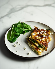 Folded omelette stuffed with spinach and feta on the white plate with spinach leaves on back. Healthy and dietary breakfast dish. Minimalistic food photo. White marble background. 