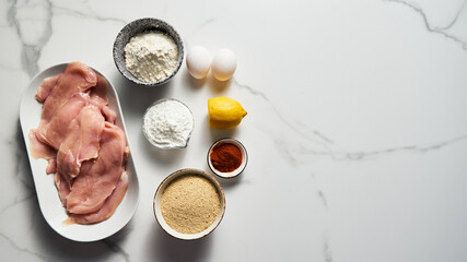 Raw Chicken fillet cut into schnitzel on an oval platter on a white marble kitchen countertop. Preparing for cooking baked chicken schnitzel. Other ingredients near