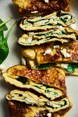 Close up of sliced folded omelette stuffed with spinach and feta on the white plate with spinach leaves on back. Healthy and dietary breakfast dish. Minimalistic food. White marble background. 