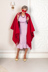 A fashionable elderly lady in a pink dress and a large scarf is draped over her shoulders is posiang against a white background.