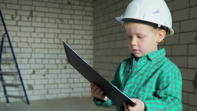 The boy in his dream became a foreman at a construction site and studies documents with room plans. A little boy of 5 years old in a white helmet and a green shirt at a construction site. 4k footage
