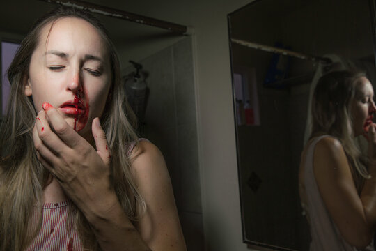 This image shows an injured young woman holding her face and closing her eyes as blood runs down her face from her nose. 