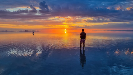Man sillhoutte walking into sunset of lake Bonneville Salt Flats, Wendover, Western Utah, USA, America. Beautiful summits of Silver Island Mountain range reflecting in water surface, Great Salt Lake