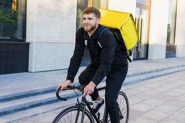 A smiling male courier rides a bicycle along a modern building in the city. A delivery man with a yellow backpack