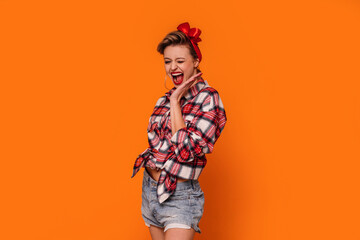 Fashion photo of happy, beautiful woman in short jeans and checkered shirt. Girl laughing, posing on orange studio background.