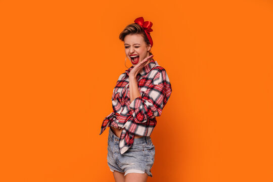 Fashion photo of happy, beautiful woman in short jeans and checkered shirt. Girl laughing, posing on orange studio background.