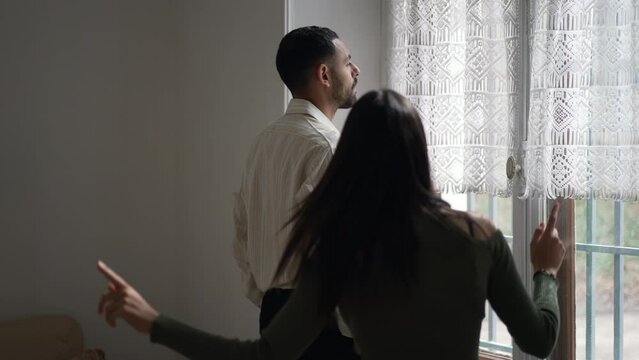 Boyfriend in silence while girlfriend makes of fun of his calm demeanor. Back of woman gesturing to man who stands by window at home
