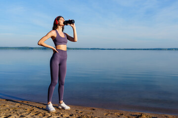 A young woman in a tracksuit drinks water after a running workout on the beach