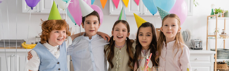positive group of children singing happy birthday song next to cake with candles and balloons,...