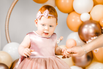 Fototapeta na wymiar Little redhead baby girl celebrates first birthday anniversary. 1 year family baloons party. Professional photoshoot. Photo studio. Cute adorable red hair kid. Red holiday dress. Children's home room