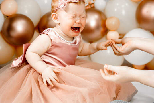 Little crying unhappy redhead baby girl celebrates first birthday anniversary. 1 year family party Professional photoshoot in photo studio. Cute adorable red hair kid in pink dress.Home children room