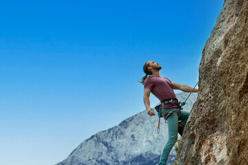 Active young man climbing on height vertical rock against blue sky. Gorgeous confident male climber looking up and overcoming a difficult route on cliff