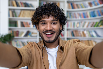 Young Hindu student programmer inside academic library taking selfie photo and talking on video call, man with curly hair smiling and looking at camera.
