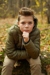 A cute stylish guy sits in the autumn forest propping up his chin with his hand and looks into the...