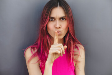 Fototapeta premium Annoyed pink hair woman showing silence sign with her index finger. Pretty female with big dark eyes frowning her eyebrows with displeasure asking to be quiet isolated against grey wall. Hush sign.