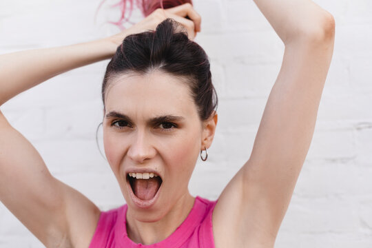 Portrait of young pink hair woman in pink top, plays with hair, arms raised up, photo on light brick background. Girl make high ponytail and screaming. Happy attractive woman braids her hair.