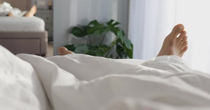 Pedestal shot unrecognizable male feet on bed in bedroom in morning. Two bare feet of man sleeping on white cotton bed clothes stick out of blanket on sunny day.