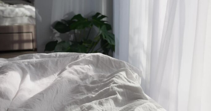 Light wind breeze in bedroom window with light tulle curtain sunny day. Beautiful shadows on unmade bed with cotton linen clothes on warm and cozy spring or summer early morning.