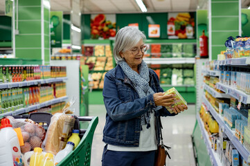 Senior Smiling Woman with Eyeglasses Pushing a Shopping Cart in the Supermarket Making Purchases -...
