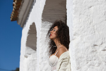 Latin woman with dark, curly hair and middle-aged dressed in jeans and white lace top is leaning on a column of a Mediterranean style house with white painted wall. The woman is on holiday
