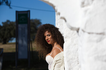 Latin woman with dark, curly hair and middle-aged dressed in jeans and white lace top is leaning on a column of a Mediterranean style house with white painted wall. The woman is on holiday
