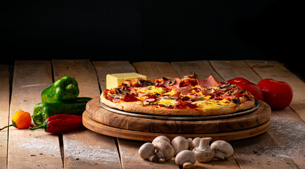 Pizza surrounded by peppers, tomatoes, mushrooms and a block of mozzarella cheese on a rustic wooden table against a black background.