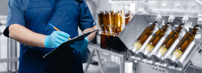 Industrial worker inspecting beer production line with glass bottles in of brewery factory with...