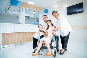 Asian family with grandmother, pregnancy woman, man and daughter stay in front of registration counter in hospital and they look at camera with happiness.