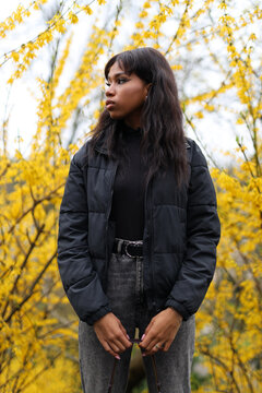 A lonely young African woman stands in a park against the backdrop of yellow bushes.
