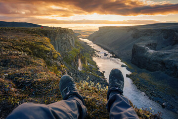 Legs of hiker man sitting on edge of mountain with waterfall flowing in canyon on Icelandic Highlands in the evening at Sigoldugljufur