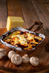 Lasagna or pasta gratin baked in an aluminum tray surrounded by mushrooms and mozzarella cheese on a rustic wooden table.