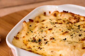 Baked lasagna or pasta gratin in a white ceramic tray surrounded by mushrooms on a rustic wooden table on a black background