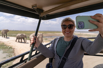 Blonde woman tourist on safari takes a selfie from the vehicle as a herd of elephants passes by © MelissaMN