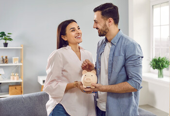 Smiling, happy young family together put coins in piggy bank to save money. Married couple are...