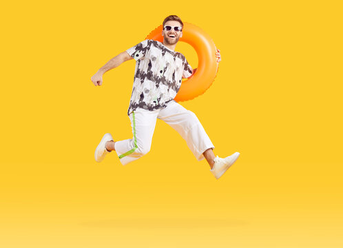 Full body photo of a young man in sunglasses holding rubber ring and jumping isolated on studio yellow background. Funny tourist is going on summer holiday trip. Vacation and travel concept.