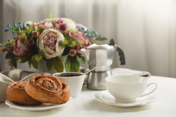 Still life breakfast in a cozy living room. 
Delicious bun, white cup and saucer and geyser coffee maker.