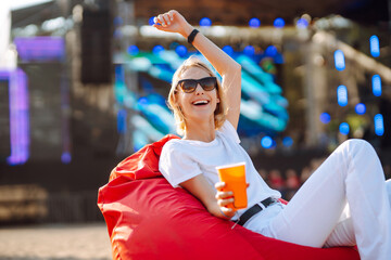 Smiling young woman drinking beer and relaxing at music festival. Happy woman is sitting on a soft pouf and relaxing while listening to a music festival. Beach party.