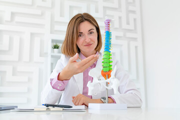 Young attractive female doctor orthopedist demonstrating the problem on spine bone model on the desk in her workplace