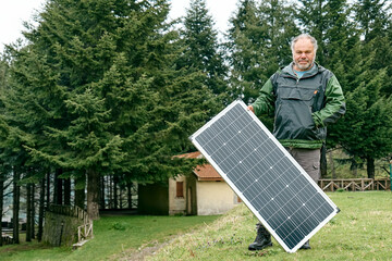 Middle aged bearded man holding solar panel near house in rural zone. Alternative energy, renewable...