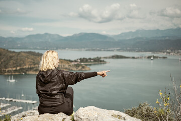 Fototapeta na wymiar Tourist, adult blonde woman admiring the view over the bay and marina in the Aegean sea, travel and hiking with adventure, healthy lifestyle outdoors, mediterranean sea nature, cloudy spring day