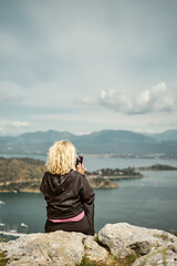 Fototapeta premium blonde woman sits on a high rock overlooking the marina and the bay in the Aegean Sea on a cloudy day and shoots a video on a smartphone. Vertical frame, travel and vacation concept