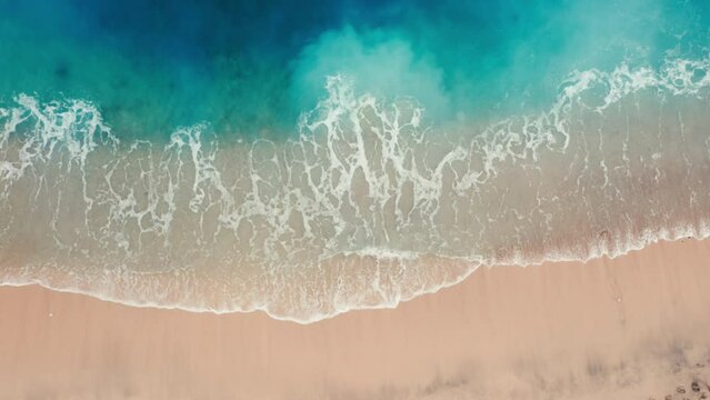 Aerial top view of beautiful seamless never ending footage while turquoise blue sea waves breaking on sandy coastline. Aerial shot of golden beach with small stones meeting ocean water and foam.