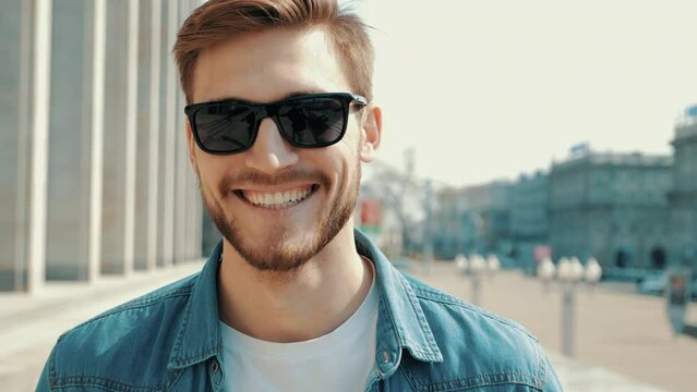 Portrait of handsome smiling stylish hipster lumbersexual model. Man dressed in jeans jacket clothes. Fashion male posing on the street background in sunglasses