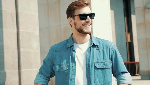 Portrait of handsome smiling stylish hipster lumbersexual model. Man dressed in jeans jacket clothes. Fashion male posing on the street background in sunglasses