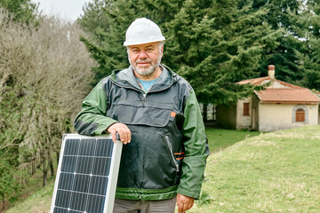 Middle aged bearded technician with voltaic solar panel near house in rural zone. Stand-alone photovoltaic solar panel system. Renewable ecological cheap green energy production. Energy independence.