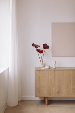 Vase with Anthurium flowers and ornaments on a wooden sideboard with blank artwork on the wall
