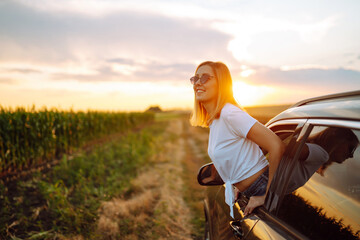 Fototapeta na wymiar Lifestyle! Happy woman enjoys sunset views from the car window. Young tourist woman rests and leans out of the car window, enjoys the trip. Travel concept, vacation. Towards adventure.