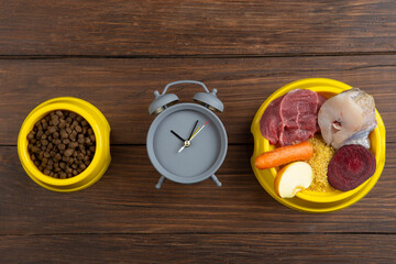 Pet care and diet concept. Bowl of dry food and raw food with meat, fish and vegetables on wooden background. Feed on time concept
