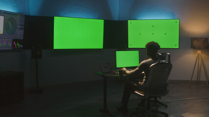 African American man works on computer in studio with equipment. Multiple chroma key big screen on...